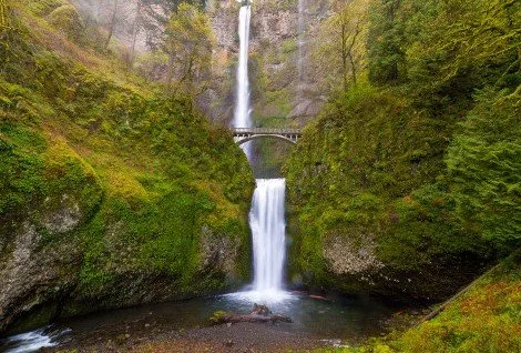 Scenic view of a cascading waterfall with a bridge, surrounded by lush greenery and steep cliffs.