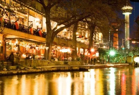 Night view of a lively riverside promenade with illuminated buildings and reflection in water, featuring a tall tower.
