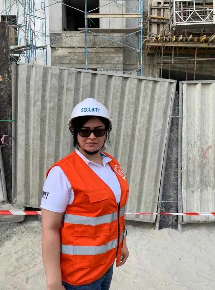 Security personnel in an orange vest and hard hat on a construction site with scaffolding and fencing in the background.