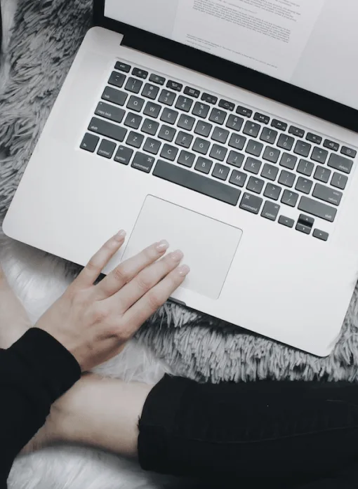 Person with laptop on lap, typing on keyboard with document on screen, sitting on gray faux fur rug.