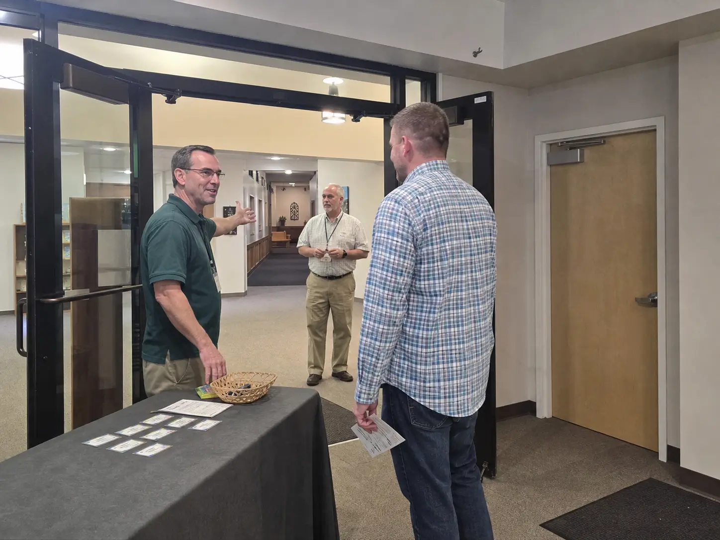 A male church greeter welcomes a visitor at a staffed entrance while another usher monitors the lobby.